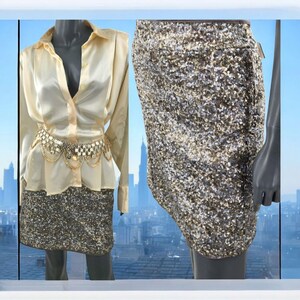 May include: A cream-coloured button-down shirt paired with a sequined skirt. The skirt is covered in small, shimmering sequins in shades of silver, gold, and bronze. A gold chain belt accents the waist. The outfit is displayed on a mannequin.