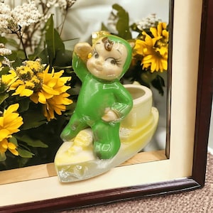 May include: A green ceramic figurine of a little person with horns, wearing a green suit, standing on a yellow boat-shaped planter. The planter has a white interior.
