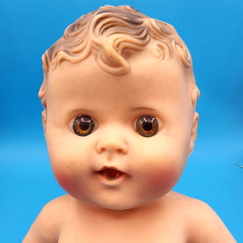 Puede incluir: Una mu&ntilde;eca beb&eacute; vintage con una expresi&oacute;n serena. La mu&ntilde;eca tiene ojos marrones, mejillas rosadas y cabello rizado casta&ntilde;o claro. El tono de piel de la mu&ntilde;eca es melocot&oacute;n p&aacute;lido, sobre un fondo azul.
