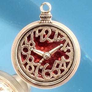 Puede incluir: Colgante plateado con diseño de esfera de reloj. La esfera tiene un fondo rojo con números y manecillas plateadas. El colgante tiene un lazo en la parte superior para sujetarlo a una cadena o pulsera. El diseño es intrincado y detallado.