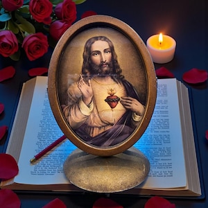 May include: An oval-framed image of Jesus Christ, featuring a detailed depiction of his face and a sacred heart. The image is set on a brass stand, placed in front of an open book, with a lit candle and red rose petals scattered around.