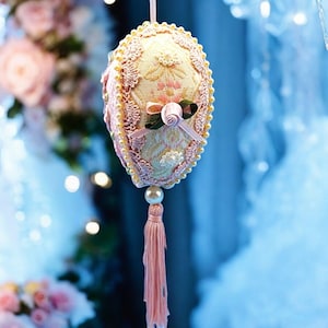 May include: A pink and white fabric Easter egg ornament with a pearl trim and a pink tassel. The egg is decorated with a floral pattern and a pink bow.