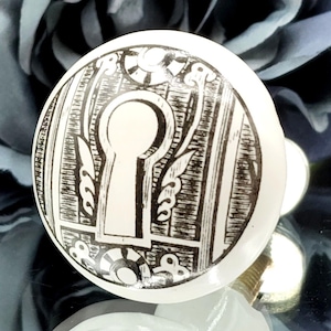 Puede incluir: Pomo de armario de cerámica blanca con diseño de cerradura negra. El pomo redondo tiene un tornillo de latón. El diseño de la cerradura está rodeado de patrones florales y geométricos decorativos.