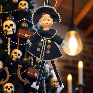 May include: A vintage-style Christmas tree ornament featuring a doll dressed in a black coat with gold trim and buttons, and a matching hat. The doll has a painted face and is hanging on a Christmas tree decorated with skulls and other ornaments.