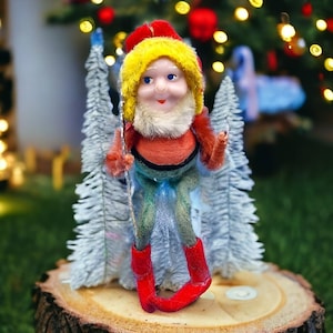 May include: A vintage Christmas elf figurine with a red hat and boots, yellow hair, and a white beard. The elf is wearing an orange shirt and green pants. It is standing in front of two small white Christmas trees on a wooden base.