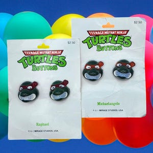 May include: Two sets of Teenage Mutant Ninja Turtles buttons. Each set includes two buttons featuring the heads of Raphael and Michelangelo. The buttons are green with red masks and white eyes. The packaging is white with the Teenage Mutant Ninja Turtles logo and the text "Teenage Mutant Ninja Turtles Buttons".