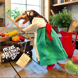 Puede incluir: Una muñeca de tela de bruja de cocina con nariz larga, que lleva un pañuelo oscuro con flores, una blusa blanca y una falda verde con una faja roja. La muñeca sostiene un palo de escoba de madera y tiene una etiqueta que dice "KITCHEN WITCH" y "MAKE YOUR OWN MAGIC".