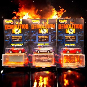 May include: Three Hot Wheels die-cast cars from the movie "Demolition Man" are displayed in their original packaging. The cars are a red Corvette Sting Ray, a silver Oldsmobile Aurora, and a red Pontiac Banshee. Each car is in a clear plastic case.