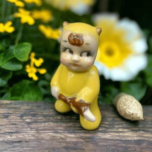 May include: A small yellow ceramic figurine of a baby wearing a yellow jumpsuit and holding a brown stick. The figurine has brown hair and large eyes.