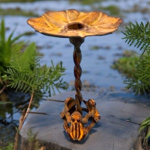 May include: A decorative metal bird bath with a yellow, leaf-shaped bowl and a twisted stem. The base features a frog design. The bird bath is made of metal and has a rustic, weathered appearance. The bird bath is designed for outdoor use.
