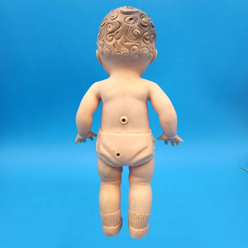 Puede incluir: Una mu&ntilde;eca beb&eacute; vintage, vista desde atr&aacute;s, con cabello moldeado detallado y un pa&ntilde;al. La mu&ntilde;eca est&aacute; hecha de un material color carne, con agujeros visibles en la espalda. Las piernas de la mu&ntilde;eca tienen calcetines moldeados.