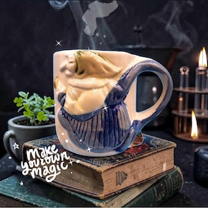 May include: A ceramic mug featuring a raised, sculpted figure of a witch in a blue dress and green hood. The mug has a curved handle and sits atop a stack of vintage books. The text "make your own magic" is written in white script on the books.