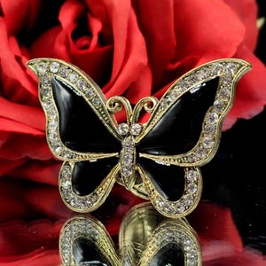 Peut inclure: Broche papillon dorée avec des ailes en émail noir et des accents de cristaux transparents. La broche est posée sur un fond rouge flou, créant un contraste saisissant. Les ailes du papillon sont bordées de cristaux étincelants.