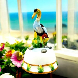 May include: A white ceramic bell with a blue and black bird figurine on top. The bird is standing on a bed of green leaves and yellow flowers. The bell has a gold trim and a white background.