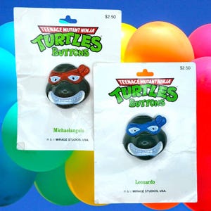 Op de afbeelding: Twee Teenage Mutant Ninja Turtles-knopen, één met een rood masker en de andere met een blauw masker. De knopen zitten op witte kaarten met de tekst "TURTLES BUTTONS" in groen en de naam van het personage. De achtergrond bevat kleurrijke ballonnen.
