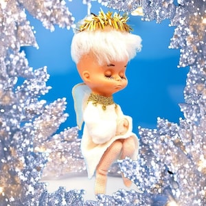 Peut inclure: Figurine d'ange vintage avec une robe blanche, des détails dorés et les yeux fermés. L'ange a des cheveux blancs et de petites ailes. La figurine est encadrée par une bordure décorative argentée scintillante, sur fond bleu.