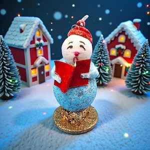 Pode incluir: Um enfeite de Natal extravagante com um boneco de neve cantando de um livro vermelho. O boneco de neve tem um chapéu vermelho e um corpo azul brilhante, sobre uma base dourada. Pequenas casas vermelhas e árvores de Natal completam a cena festiva.