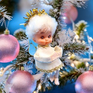 Peut inclure: Ornement de Noël en forme d'ange. L'ange porte une robe blanche avec des bordures dorées, une chevelure blanche duveteuse et une couronne dorée. Il a des ailes bleues et est suspendu à un sapin de Noël avec des boules roses.