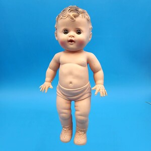 Puede incluir: Una mu&ntilde;eca beb&eacute; vintage con piel clara, ojos marrones y la boca ligeramente abierta. La mu&ntilde;eca tiene cabello casta&ntilde;o rizado y lleva un pa&ntilde;al y zapatos. Las mejillas de la mu&ntilde;eca est&aacute;n sonrojadas y est&aacute; posada sobre un fondo azul.