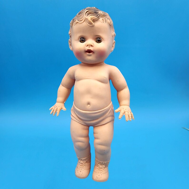 Puede incluir: Una mu&ntilde;eca beb&eacute; vintage con piel clara, ojos marrones y la boca ligeramente abierta. La mu&ntilde;eca tiene cabello casta&ntilde;o rizado y lleva un pa&ntilde;al y zapatos. Las mejillas de la mu&ntilde;eca est&aacute;n sonrojadas y est&aacute; posada sobre un fondo azul.