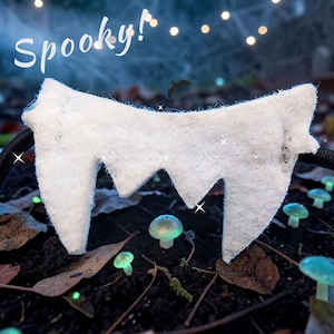 May include: A white felt banner with a jagged, tooth-like bottom edge featuring the word Spooky! in white script text. The banner is positioned above small, glowing, light blue mushrooms on dark soil with scattered brown leaves and bokeh lights.