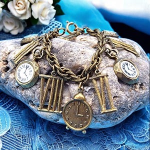 May include: A vintage-style charm bracelet with gold-toned charms. The bracelet features clock-themed charms, including miniature pocket watches and Roman numeral designs. The bracelet is displayed on a gray stone against a blue background.