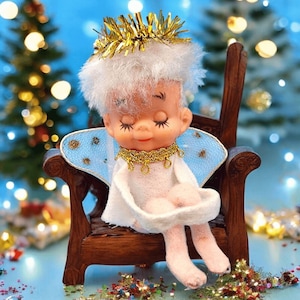Peut inclure: Une poupée ange vintage aux yeux fermés, portant un halo et un collier dorés, assise dans une chaise en bois miniature. La poupée a une robe et des ailes blanches, avec des jambes roses et un panier blanc. Le fond est bleu doux avec des lumières de Noël floues.