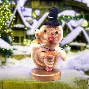 Puede incluir: Una figura caprichosa de un muñeco de nieve de peluche con un sombrero negro, gafas redondas y una nariz roja. El muñeco de nieve sostiene un pequeño tambor naranja con baquetas negras. Está montado sobre una base redonda de madera, con un fondo de pueblo nevado.