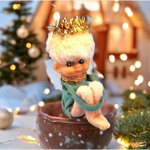 Peut inclure: Une poupée de lutin de Noël fantaisiste avec une tête duveteuse de couleur claire, vêtue d'une tenue verte et d'accents dorés. Le lutin est assis dans un bol marron, avec une couronne de guirlande dorée et des ailes blanches. L'arrière-plan présente un sapin de Noël et des lumières.