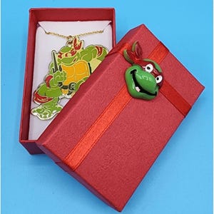 May include: A gold-colored necklace featuring a green, yellow, and red Teenage Mutant Ninja Turtle pendant, presented in an open red gift box. The box is adorned with a red ribbon and a turtle head embellishment. The necklace is displayed on a white card.