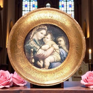 May include: A round, gold-framed painting depicting a mother holding a child, with another figure to the side. The painting has a dark background and is set on a wooden stand. Pink roses are in the foreground.