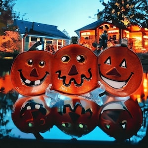 Puede incluir: Un trío de calabazas naranjas sonrientes con rasgos faciales negros y tallos verdes. Las calabazas se reflejan en el agua, con un fondo borroso de casas y árboles, creando una escena festiva de Halloween.