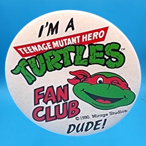 May include: A round, off-white button featuring the Teenage Mutant Ninja Turtles. The button has the words "I'M A TEENAGE MUTANT HERO TURTLES FAN CLUB DUDE!" in black, red, and green. A smiling turtle head with a red mask is also visible.