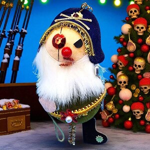 May include: A quirky Christmas decoration in the form of a gnome with a blue hat, white beard, and a red nose. The gnome is embellished with gold trim and a red jewel eye. A Christmas tree decorated with skull ornaments is in the background.