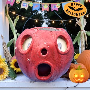 May include: A red, round Halloween pumpkin decoration with large eye and mouth openings. The pumpkin has a metal handle and is surrounded by other pumpkins and sunflowers. The image includes a "Happy Halloween" sign.