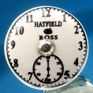 Puede incluir: Una esfera de reloj blanca con números negros y las palabras "HATFIELD ROSS". Un dial más pequeño está en la parte inferior. La esfera del reloj tiene un botón de metal plateado en el centro.
