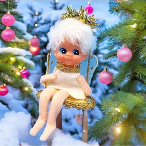 May include: A doll with white hair and blue eyes sits on a gold-trimmed chair. The doll wears a white dress and a gold collar. Pink ornaments and a Christmas tree are in the background, creating a festive holiday scene.