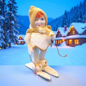 Puede incluir: Adorno navideño vintage de una muñeca esquiadora. La muñeca tiene una capucha y capa amarillas, con cuerpo y esquís blancos. Sostiene cuerdas rojas, con un fondo de casas y árboles nevados.