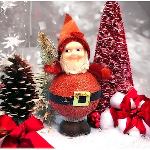 Op de afbeelding: Een kerstkabouterfiguur met een rode glitterende body, zwarte riem en een gouden gesp. De kabouter heeft een rode hoed, witte baard en blauwe ogen. De scène bevat een rode kerstboom, dennenappel en een rode strik.