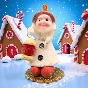 May include: A vintage Christmas ornament featuring a smiling gnome holding a lantern. The gnome wears a white coat with black buttons, red feet, and a brown hat. Gingerbread houses with candy cane accents are in the background, creating a festive holiday scene.