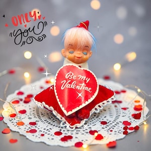 Puede incluir: Figura de San Valentín de una muñeca sosteniendo un letrero rojo en forma de corazón que dice "Be my Valentine". La muñeca tiene los ojos cerrados, cabello blanco y un sombrero rojo. El letrero está sobre una almohada roja, decorada con un mantel individual blanco y confeti de corazón rojo.