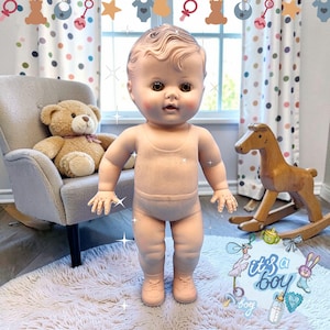 May include: A vinyl baby doll with moulded hair, brown eyes, and rosy cheeks, wearing a light pink ribbed one-piece outfit and matching shoes. The doll stands upright, featuring articulated limbs and a detailed facial expression.