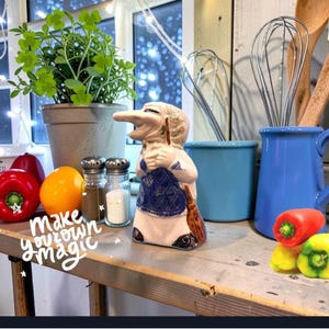 May include: A ceramic troll figurine with a long nose, wearing a blue apron and holding a broom, positioned between salt and pepper shakers, a blue pitcher with metal whisks, and colorful bell peppers with the text make your own magic.