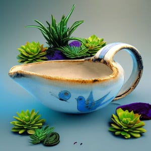 May include: A blue and white ceramic gravy boat with a blue bird and butterfly design. The boat has a handle and is sitting on a table with green plants.