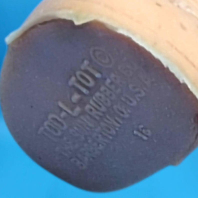 Puede incluir: Primer plano de la base de un juguete de goma vintage. La base es de color p&uacute;rpura oscuro con el texto "TOO-L-TOT" y "SUN RUBBER" impreso. El juguete es de color marr&oacute;n claro y tiene forma redondeada.