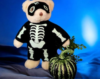 Puede incluir: Un oso de peluche blanco con un disfraz de esqueleto negro y una máscara negra para los ojos. El oso sostiene una calabaza verde y negra.