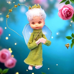 Puede incluir: Una muñeca caprichosa vestida de ángel, con un vestido de punto verde y una corona dorada. La muñeca tiene cabello blanco, alas azules y guantes rosas. Una varita dorada en forma de estrella se extiende de la mano de la muñeca. Rosas rosas y hojas verdes rodean la muñeca sobre un fondo azul.