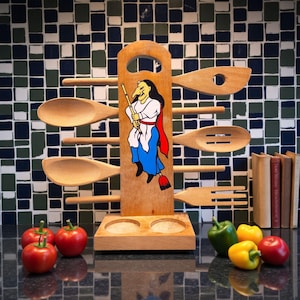 May include: Wooden kitchen utensil holder with a painted witch design. Includes wooden spoons, a slotted spoon, and a fork. The holder has two circular openings at the base. Red tomatoes and yellow and green bell peppers are in the foreground.