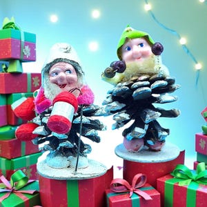 Puede incluir: Dos figuras caprichosas de gnomos navideños hechas de piñas, cada una con una cara sonriente y vestimenta festiva. Un gnomo sostiene una linterna roja y blanca, el otro un gorro verde. Se asientan sobre cajas de regalo rojas y blancas, rodeadas de más regalos envueltos.