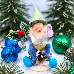 May include: A whimsical Christmas gnome figurine with a green hat, white beard, and blue pants. The gnome is holding a sprig of holly with red berries. The background includes blue and green Christmas ornaments and evergreen branches.